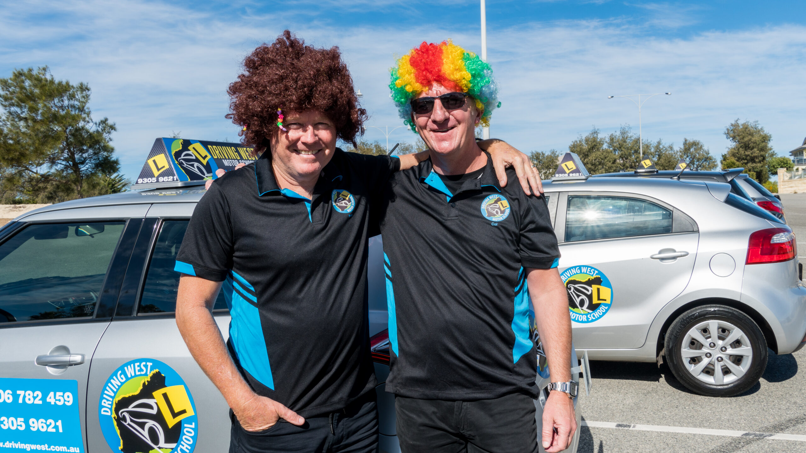 Driving West colourful wigs photoshoot 2019 Two Driving West team members wearing colourful wigs and smiling beside learner cars during the 2019 photoshoot